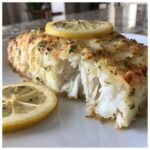 Close-up of flaky, oven-baked chilean sea bass garnished with lemon slices and parsley.