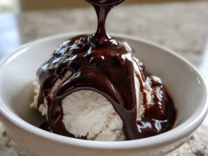 Close-up of thick, glossy chocolate sauce for ice cream being poured over a scoop of vanilla ice cream in a white bowl.