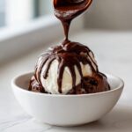 A spoonful of rich, homemade chocolate sauce for ice cream is being poured over vanilla and chocolate ice cream scoops in a white bowl.