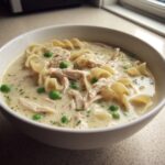 Close-up of a white bowl filled with creamy chicken noodle soup, featuring shredded chicken, egg noodles, and bright green peas.
