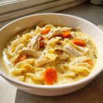 A close-up of a bowl of creamy chicken noodle soup crock pot, featuring egg noodles, shredded chicken, and carrots, seasoned with pepper.