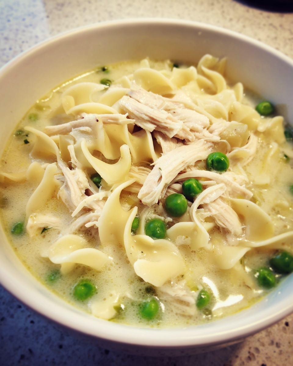 Close-up of a white bowl filled with creamy chicken noodle soup, featuring egg noodles, shredded chicken, and bright green peas.