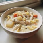 Close-up of a white bowl filled with creamy chicken noodle soup with potatoes and carrots.