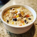 Close-up of a white bowl filled with creamy chicken tortilla soup crock pot, topped with shredded chicken, corn, and black beans.