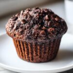 A close-up photo of a rich, dark double chocolate muffin topped with mini chocolate chips, sitting on a white plate.