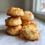 A close-up stack of golden brown, chewy coconut cookies resting on a white marble surface.