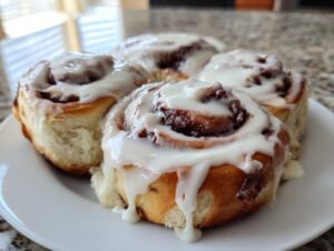 Close-up of four gooey cinnamon rolls generously topped with dripping white cream cheese icing on a white plate.