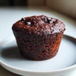 Close-up of a single, dark, moist healthy chocolate muffin topped with chocolate chips on a light plate.