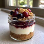A layered individual Protein Cheesecake served in a small glass jar, topped with berry compote, crumble, and fresh berries.