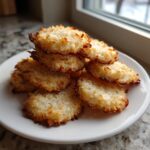 A stack of golden brown, crispy keto coconut cookies piled on a white plate near a window.