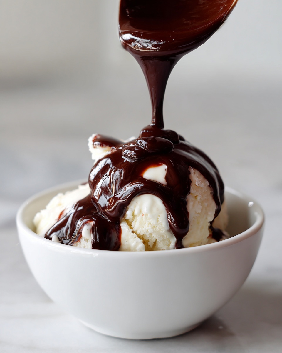 A spoonful of thick, glossy chocolate sauce for ice cream being poured over a scoop of vanilla ice cream in a white bowl.