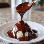 A spoonful of thick, glossy chocolate sauce is being poured over a scoop of vanilla ice cream.