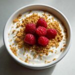 Overhead view of a creamy protein cheesecake bowl topped with crushed graham crackers and fresh raspberries.
