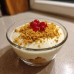 A serving of creamy protein cheesecake pudding topped with graham cracker crumbs and fresh red currants.