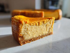 A close-up slice of rich pumpkin protein cheesecake with a graham cracker crust, sitting on a white counter.