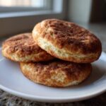 Close-up of three soft, round snickerdoodle cookies stacked on a white plate, coated heavily in cinnamon sugar.