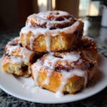 A stack of three soft sourdough cinnamon rolls generously drizzled with white cream cheese icing on a white plate.