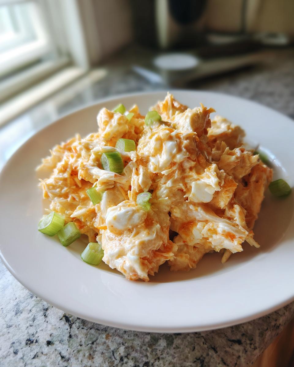 A mound of creamy, orange-tinted spicy crab salad garnished with chopped green onions on a white plate.