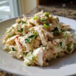 Close-up of a mound of creamy, shredded spicy crab salad topped with chopped green onions on a white plate.