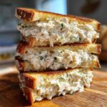 Three stacked halves of a rich crab salad sandwich overflowing with creamy filling on toasted bread.