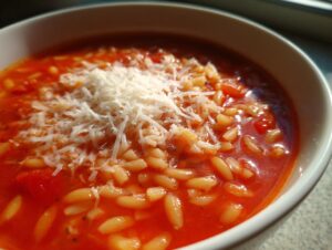 A close-up shot of a bowl of vibrant tomato pastina soup topped generously with grated Parmesan cheese.