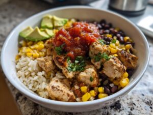 A close-up of a white bowl filled with a delicious chicken burrito bowl, featuring seasoned chicken, brown rice, black beans, corn, salsa, and avocado slices.