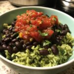 A close-up of a vibrant black bean and rice bowl featuring green cilantro rice, black beans, and a topping of fresh tomato salsa.