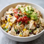 Close-up of a white bowl filled with a delicious chicken burrito bowl, featuring rice, seasoned chicken, corn, black beans, salsa, and avocado.