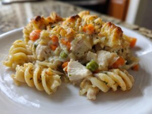 A close-up serving of creamy chicken pot pie pasta featuring fusilli, chicken chunks, carrots, and peas, topped with a golden, baked crust.