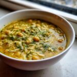 Close-up of a white bowl filled with yellow chickpea soup, orzo pasta, and fresh dill.