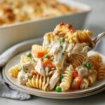 A spoonful of creamy chicken pot pie pasta, featuring rotini, chicken chunks, peas, and carrots, lifted from a plate.