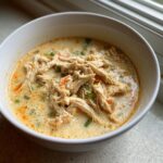 A close-up of a white bowl filled with creamy slow cooker buffalo white chicken chili, topped with shredded chicken and visible vegetable pieces.
