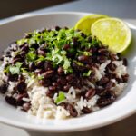 A close-up bowl of white rice topped with seasoned black beans and fresh cilantro, served with lime wedges.