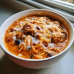 A close-up of a white bowl filled with creamy, orange-red slow cooker buffalo chicken chili, topped with shredded cheese and a drizzle of white sauce.