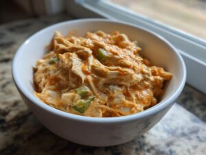 A close-up of shredded chicken coated in orange buffalo sauce and mixed with green peppers in a white bowl, representing slow cooker buffalo white chicken chili.