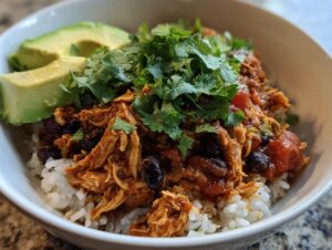 A close-up of a finished slow cooker chicken burrito bowl featuring shredded chicken, black beans, rice, avocado, and fresh cilantro.