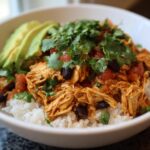 Close-up of a white bowl filled with rice, topped with shredded chicken, black beans, salsa, and fresh cilantro, served with sliced avocado.