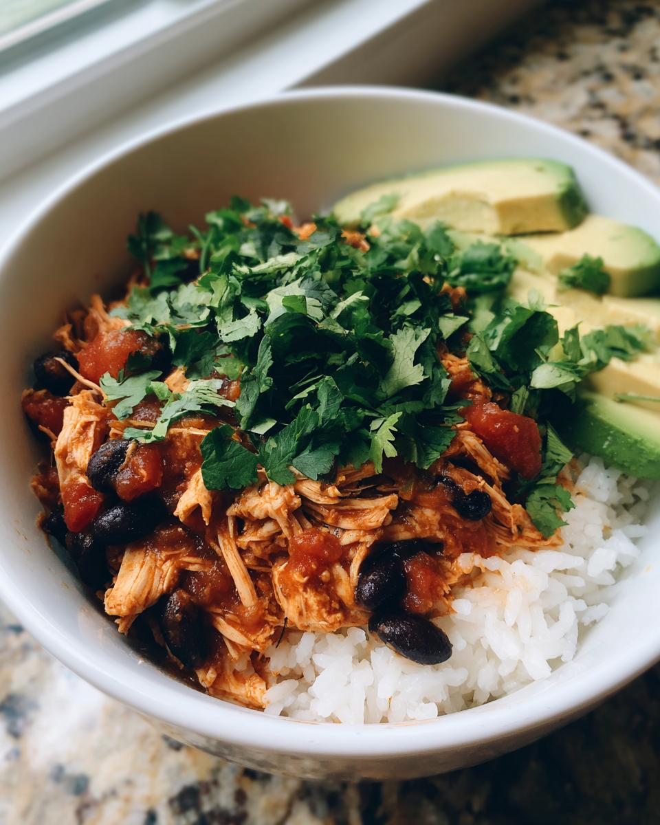 A white bowl filled with white rice topped with shredded chicken, black beans, salsa, and fresh cilantro, served with sliced avocado on the side for a slow cooker chicken burrito bowl.