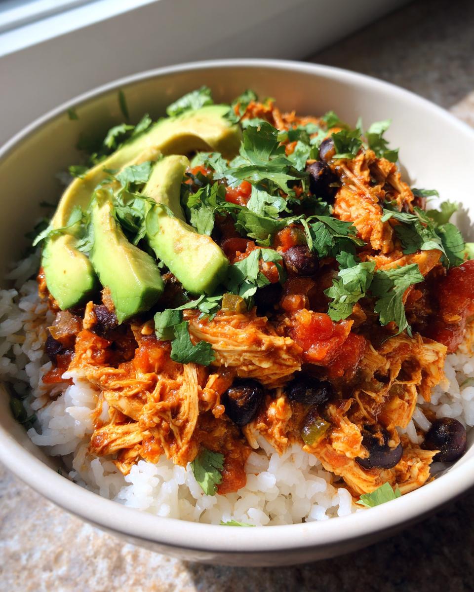 A close-up of a delicious slow cooker chicken burrito bowl served over white rice, topped with shredded chicken, black beans, avocado slices, and cilantro.