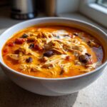 Close-up of a white bowl filled with creamy, orange slow cooker gourmet buffalo chicken chili, topped with shredded chicken and a swirl of cream.