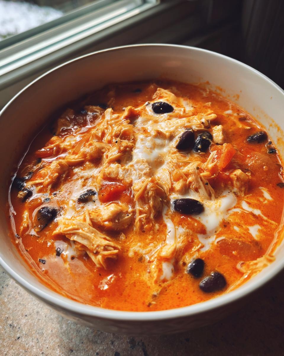 Close-up of a bowl of rich, orange slow cooker gourmet buffalo chicken chili topped with sour cream swirl and black beans.